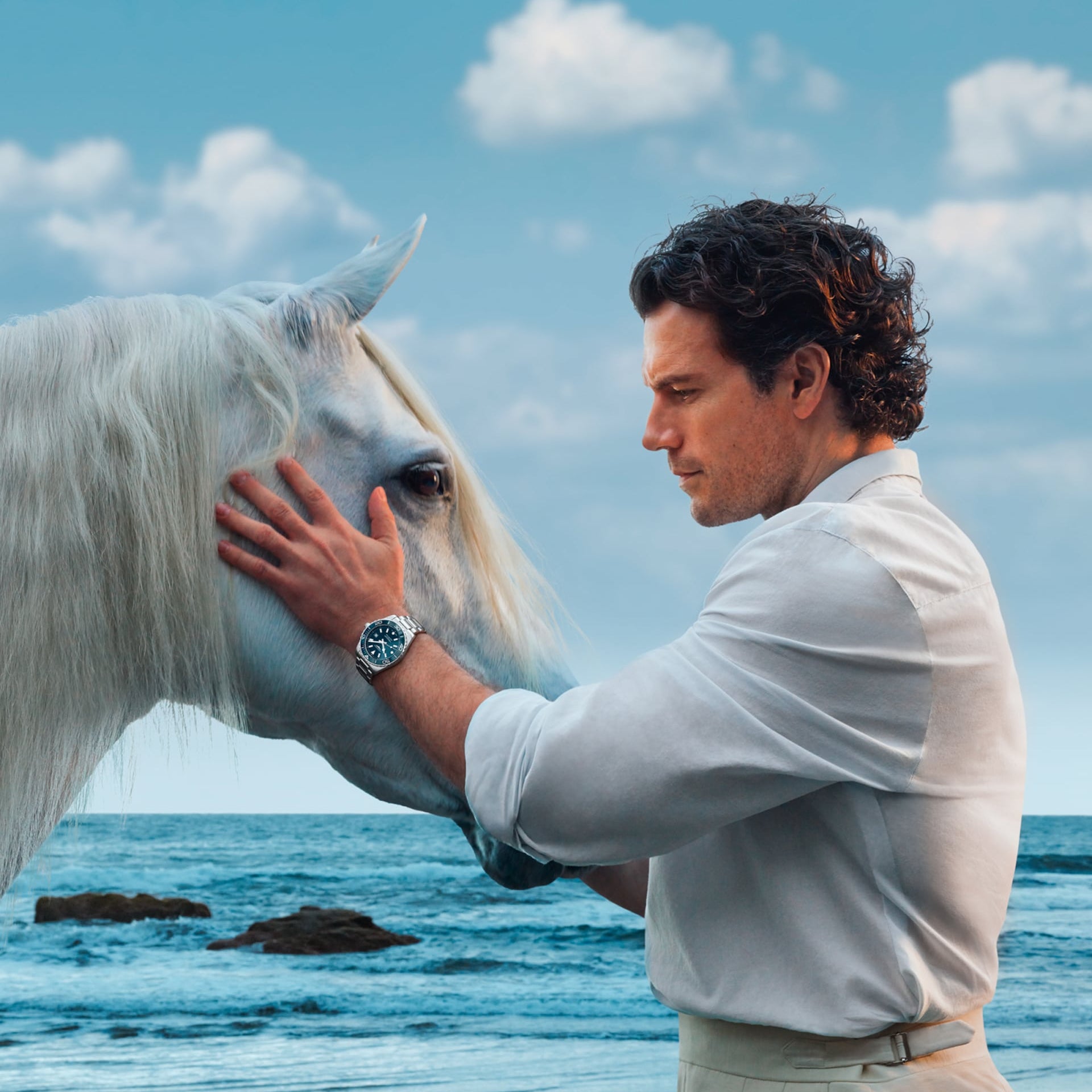 Man petting a white horse on a beach with blue sky and clouds.